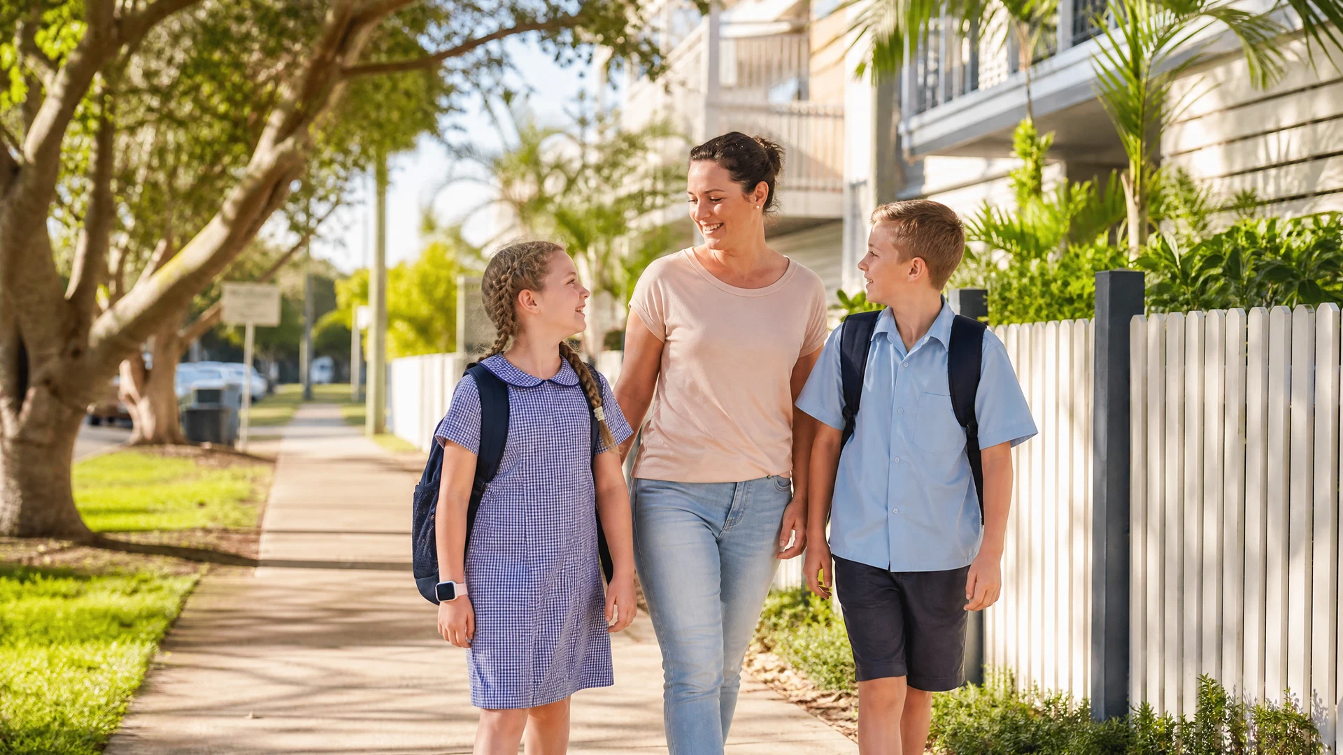 Mother and two children walking along a suburban Australian footpath on a sunny morning with school bags